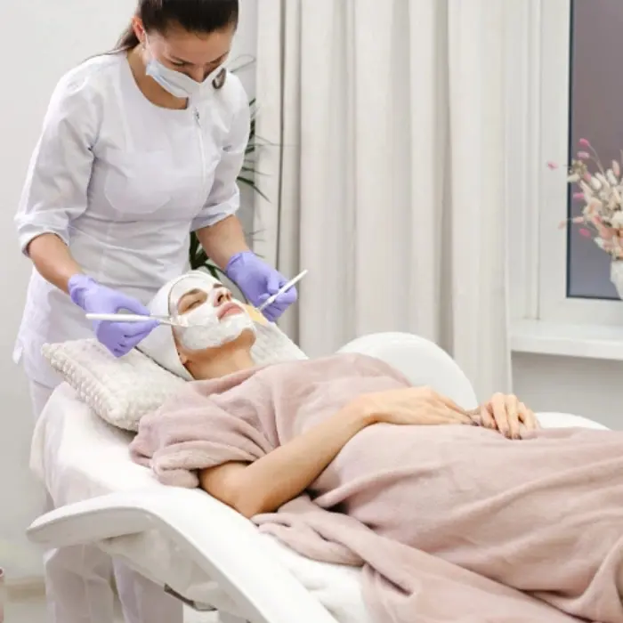 It is an image of a wellness clinic where a female professional wearing gloves is applying face mask on a female lying on treatment bed